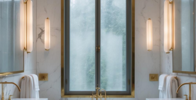Symmetrical luxury bathroom featuring white marble walls and floors, a central freestanding bathtub under a large arched window, double vanities with brass fixtures, and warm vertical wall sconces.