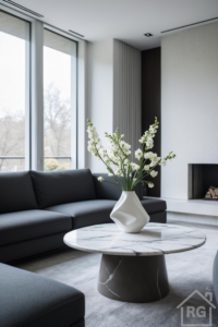 A modern minimalist living room featuring a dark grey sofa facing a round coffee table with a white marble top and dark grey base. A white sculptural vase with tall white flowers sits on the table, illuminated by large windows.