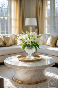 A bright, elegant living room featuring a white tufted sofa with gold pillows, centered around a round white marble coffee table with a gold base trim. A crystal vase with white lilies sits on a gold tray on the table, illuminated by sunlight from large windows with gold curtains.
