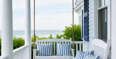 A serene coastal porch with white painted wood, a light blue ceiling, a white porch swing, a white rocking chair, blue and white striped pillows, and potted white hydrangeas, overlooking the ocean.