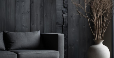 A minimalist Japandi Wabi Sabi living room featuring a dark charcoal shou sugi ban wood paneled wall, a grey fabric sofa, and a raw, dark wood coffee table. A large light ceramic vase with dried branches and a small ceramic bowl complete the serene, earthy aesthetic.