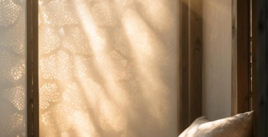 A sun-drenched Japandi-style sitting nook featuring a low wooden bench with light beige linen cushions. Golden light streams through a decorative patterned screen, casting intricate shadows on the wall.