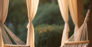 A cozy outdoor retreat featuring a comfortable beige fabric hammock with a white pillow, surrounded by flowing cream outdoor curtains and illuminated by warm string lights, set against a lush green background.