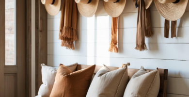 A cozy modern farmhouse entryway featuring a light wood bench adorned with beige and terracotta pillows, set against a white shiplap wall. Straw hats and scarves hang from black hooks above, with woven baskets and a jute rug on the floor, creating a warm and inviting space.