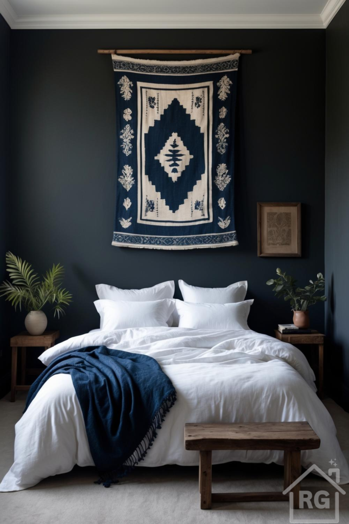 A serene dark boho bedroom featuring deep charcoal blue walls, white linen bedding, and a large blue and white woven tapestry above the bed. Rustic wooden nightstands with green plants flank the bed, and a wooden bench sits at the foot, creating a cozy and natural aesthetic.