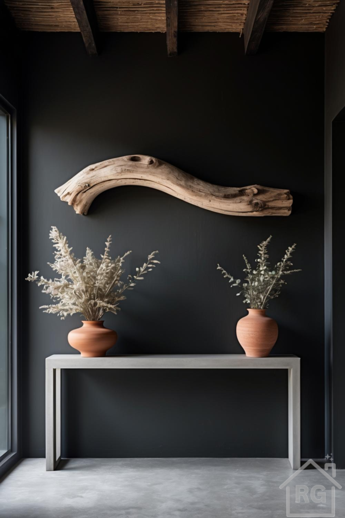A dark charcoal-painted foyer features a light grey concrete console table adorned with two terracotta vases holding dried botanicals. Above the table, a large, curved piece of light-colored driftwood serves as a striking wall art piece, complementing the rustic ceiling beams.