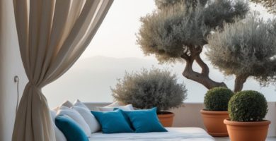 A serene Mediterranean-style patio features a luxurious outdoor daybed with white and teal pillows, framed by flowing beige curtains. Large potted olive trees and spherical shrubs in terracotta planters adorn the grey stone tiled floor, overlooking a distant mountain landscape.