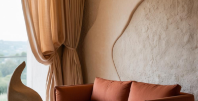 A modern living room featuring a terracotta-colored sofa, a rough-hewn stone coffee table with abstract sculptures, and a textured wall with organic shapes. Light beige curtains frame a large window, and warm cove lighting illuminates the textured wall.