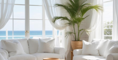 A bright and airy coastal living room with white slipcovered sofas, a light wood coffee table, and a large palm plant, overlooking a blue ocean through sheer white curtains. The room features white painted wooden floors and a natural fiber rug.