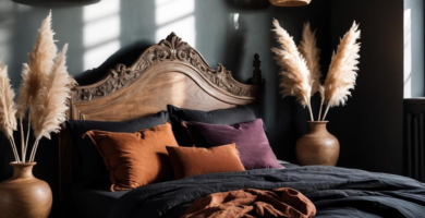 A moody dark boho bedroom featuring a carved wooden bed frame with black linen bedding, burnt orange and plum velvet pillows, and a rust-colored throw. Woven rattan pendant lights and large wooden vases with pampas grass flank the bed, set against dark charcoal walls with sunlight streaming through a window.