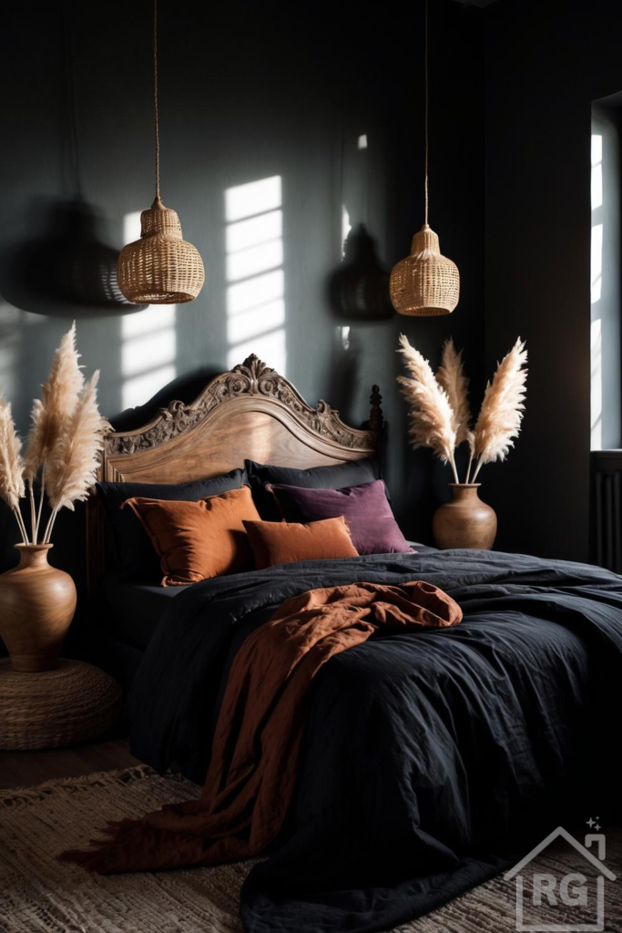 A moody dark boho bedroom featuring a carved wooden bed frame with black linen bedding, burnt orange and plum velvet pillows, and a rust-colored throw. Woven rattan pendant lights and large wooden vases with pampas grass flank the bed, set against dark charcoal walls with sunlight streaming through a window.