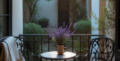 A cozy balcony features a black wrought iron bistro set with a lavender plant on the table, a beige throw blanket draped over one chair, and light-colored floor tiles, overlooking a lush green courtyard under warm sunlight.