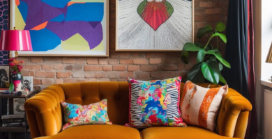 A vibrant and eclectic living room featuring an exposed brick wall, a mustard velvet sofa adorned with colorful patterned pillows, and a large geometric area rug. Two large abstract art prints hang above the sofa, complementing the urban industrial aesthetic.