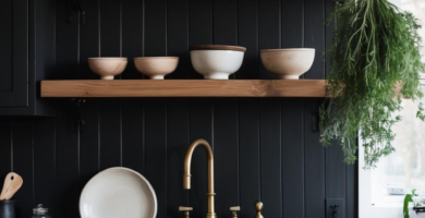 A dark boho kitchen featuring black shaker cabinets, natural wood countertops, and open shelving styled with ceramic and wooden bowls. A brass faucet and hanging dried herbs add to the rustic, organized aesthetic.