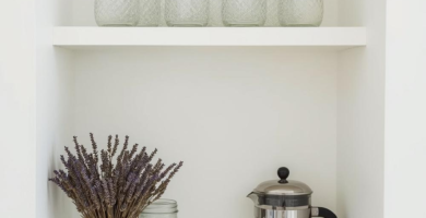 A minimalist coffee nook featuring a built-in arched white niche with two shelves. The top shelf holds several textured clear glass jars, while the bottom shelf displays a French press coffee maker, a glass jar with dried lavender, and another clear glass jar, all above a white cabinet with a wooden pull. This serene setup highlights clean design and natural elements.