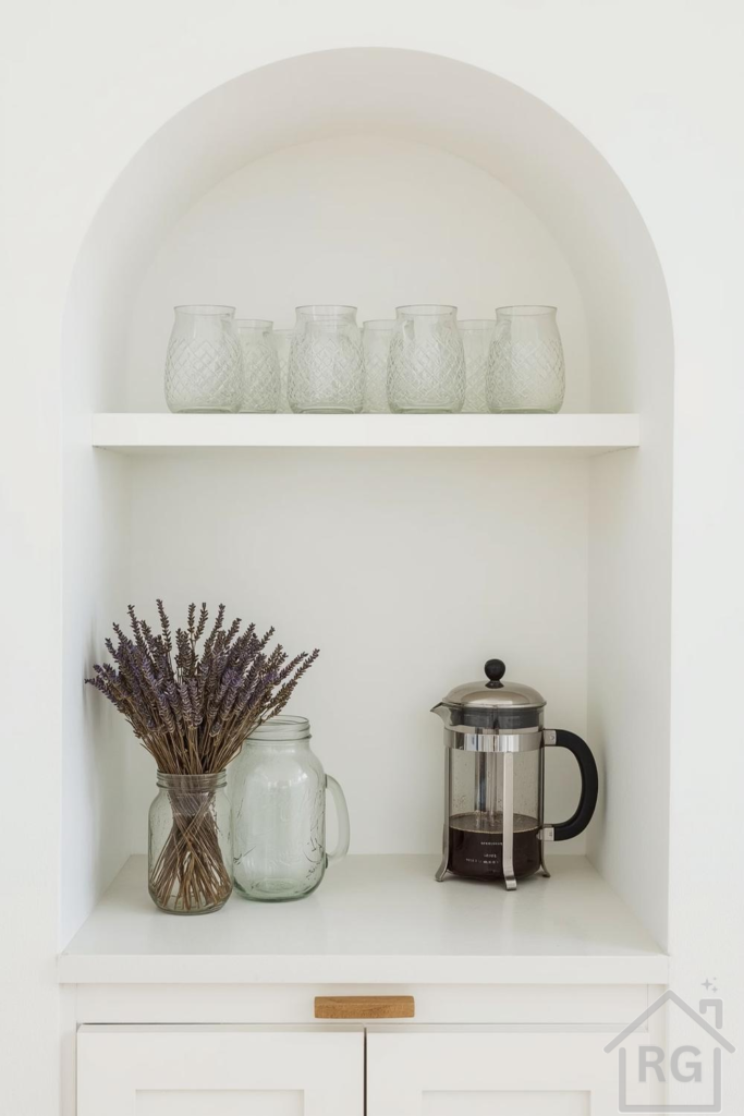 A minimalist coffee nook featuring a built-in arched white niche with two shelves. The top shelf holds several textured clear glass jars, while the bottom shelf displays a French press coffee maker, a glass jar with dried lavender, and another clear glass jar, all above a white cabinet with a wooden pull. This serene setup highlights clean design and natural elements.
