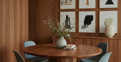 A stylish mid-century modern dining room featuring rich wood paneling, a round pedestal dining table, and four light blue chairs. The space is illuminated by a geometric pendant light and decorated with a gallery wall of abstract art prints.