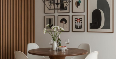 A sleek midcentury modern dining room featuring a round wooden table, four white Eames-style chairs, and a black geometric chandelier. The space is accented by a slatted wood wall panel and a gallery wall of abstract art prints, creating a warm and stylish dining room.