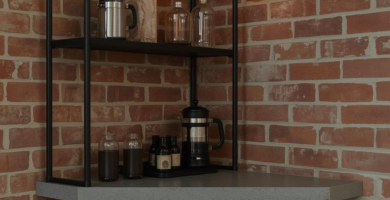A stylish industrial loft coffee corner featuring exposed red brick walls, black metal shelving, and a grey countertop. The space is illuminated by a warm Edison bulb sconce and includes two French presses, glass jars, and a wooden bar stool, creating a cozy and functional brew station.