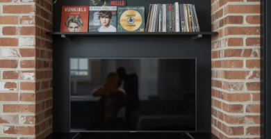 An urban industrial TV wall design integrated into an exposed brick archway. It features matte black floating shelves, a TV cabinet, a large flat-screen television, decorative records, a trailing plant, and an industrial-style wall lamp, showcasing a stylish tv wall design.