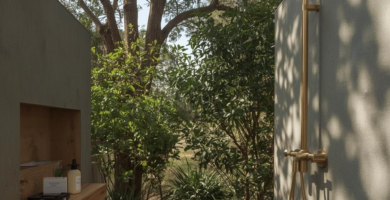 An inviting outdoor shower area featuring a brass shower fixture mounted on a grey concrete wall, a wooden slatted deck, and a wooden stool, surrounded by lush green foliage and a large tree. White pebbles line the base of the wall, adding a natural touch to this serene bathing space.