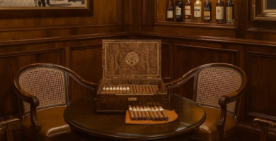 A luxurious, traditional cigar lounge featuring dark wood paneling, four brown leather armchairs, a round table with an open humidor, and built-in shelves displaying liquor bottles. A large framed black and white artwork hangs on the wall, and an ornate area rug covers the floor.