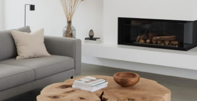 A minimalist living room featuring a large, natural wood rustic coffee table made from a tree stump. The room includes a light grey sofa, a modern black fireplace, and pampas grass in a clear vase, creating a serene and organic modern aesthetic.