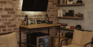 A cozy and functional small man cave featuring an exposed brick wall, concrete ceiling, and industrial-style furniture. The room includes two beige armchairs, a wooden console table with a wall-mounted TV and gaming setup, floating shelves with books and decor, a mini-fridge, and a patterned area rug. This space is designed for relaxation and entertainment, ideal for a small man cave or garage conversion.