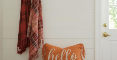 A cozy and functional mudroom entryway featuring white shiplap walls, a light wood bench, and built-in shelving. The space is decorated with fall decor including an orange "hello" pillow, plaid scarves, and wicker baskets, creating a warm and inviting atmosphere.