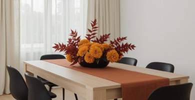 A minimalist dining room featuring a light wood table, sleek black chairs, and light beige curtains. The table is adorned with a burnt orange runner and a vibrant fall centerpiece of orange flowers and reddish-brown leaves, creating a cozy and inviting atmosphere.
