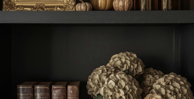A dark, moody bookshelf styled with a fall aesthetic. The shelves feature a gold-framed portrait, amber glass bottles, decorative pumpkins, vintage leather-bound books, and dried hydrangeas in a metal pitcher, creating a rich and inviting vignette.