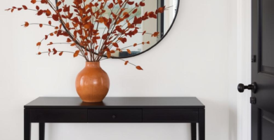 A modern and elegant entrance foyer styled with fall decor. A black console table holds a terracotta vase with rust-colored fall foliage, beneath a large round black-framed mirror, next to a black door. A natural fiber rug and brown boots complete the inviting scene.
