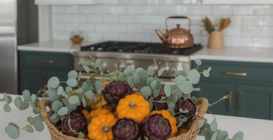 A modern farmhouse kitchen featuring white upper cabinets, deep green lower cabinets, and a white subway tile backsplash. A large woven basket filled with vibrant orange mini pumpkins, deep purple artichokes, and muted green eucalyptus branches sits on a white marble-look island, adding a touch of inviting fall decor.