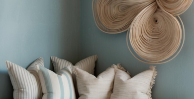 A cozy coastal-inspired daybed nook featuring a distressed white daybed frame, a striped blue and white mattress, and an assortment of throw pillows. A large, organic woven rattan wall sculpture adorns the muted blue wall above the daybed.