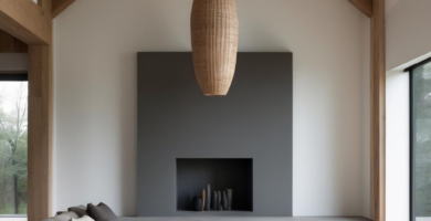 A modern Scandinavian cabin living room featuring a high vault ceiling with exposed wooden beams. The space includes a light grey sectional sofa, a dark grey minimalist fireplace, and large windows, creating a bright and serene interior.