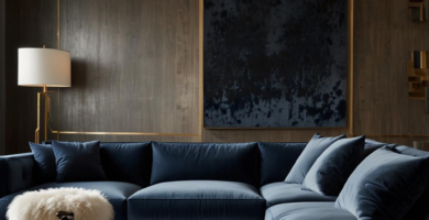 A luxurious living room featuring a large navy blue velvet sectional sofa, a dark wood round coffee table, and a light abstract rug. The walls are paneled in dark wood with brass accents, complemented by a striking gold chandelier and a tall brass floor lamp, creating a sophisticated and inviting atmosphere.
