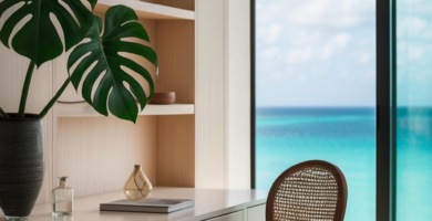 A modern coastal home office featuring a light wood built-in desk and shelving unit, a classic cane chair, and a large Monstera plant. The room offers a stunning panoramic view of a turquoise ocean and blue sky through a large window, creating a serene and inspiring workspace.