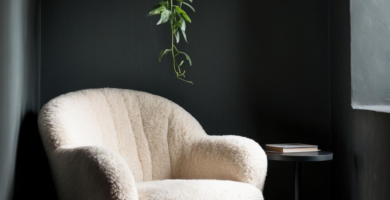 A cozy dark boho reading nook featuring a cream boucle armchair, a vintage patterned rug, and a hanging green plant. The space is set within a dark grey arched alcove with a small black side table holding books, creating an inviting and serene atmosphere.