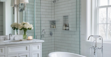 A bright and elegant transitional bathroom featuring a white freestanding bathtub, a white vanity with a marble countertop, and a glass-enclosed shower with white subway tiles. The room is illuminated by natural light from a large window with a grey Roman shade, creating a serene and luxurious atmosphere.
