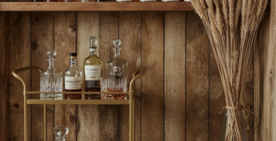 A cozy rustic home bar setup featuring a wooden plank wall, built-in whiskey shelves, and a gold bar cart laden with liquor bottles and decanters. A brown leather ottoman and a tall vase with dried wheat complete this inviting space.