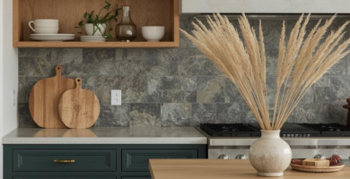A modern organic kitchen featuring deep forest green base cabinets and a matching island, complemented by light natural wood open shelving and countertops. The space includes a grey stone tile backsplash, white ceramic dishes, wood cutting boards, and a woven pendant light, creating a warm and inviting atmosphere.