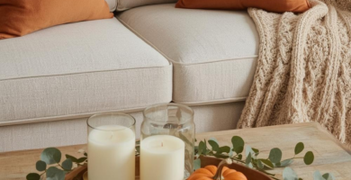 A cozy living room scene featuring a cream sofa adorned with burnt orange throw pillows and a beige cable knit blanket. A rustic wooden coffee table holds a decorative tray with white pillar candles, a small orange pumpkin, and eucalyptus stems, creating a warm fall decor ambiance.