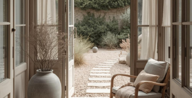 A serene sunroom or enclosed porch with light wood paneling and stone tile flooring, featuring a rattan armchair with cushions, a large ceramic vase with dried branches, and sheer curtains. The open glass door leads to a gravel patio with stepping stones and lush green foliage.