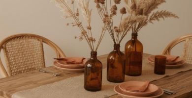 A minimalist dining room table set for fall, featuring a wooden table, rattan chairs, a burlap runner, and amber glass vases with dried pampas grass and botanicals. The table is adorned with terracotta-colored plates and linen napkins, creating a warm and inviting autumn aesthetic.